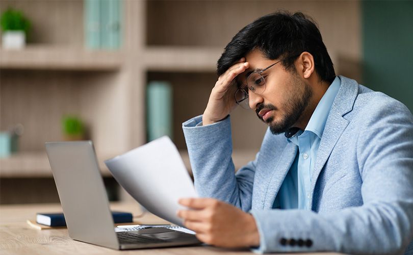 Stressed young adult sitting at a desk surrounded by bills, loan documents, and a laptop—symbolizing the emotional burden and mental strain caused by debt and financial anxiety.
